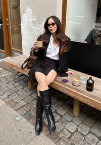 Schwarze Leder-Stiefel bis zum Knie, weißes Hemd und schwarzer Rock. Sitzt auf einer Holzbank mit einer braunen Tasche und Glaswaren auf der Oberfläche.