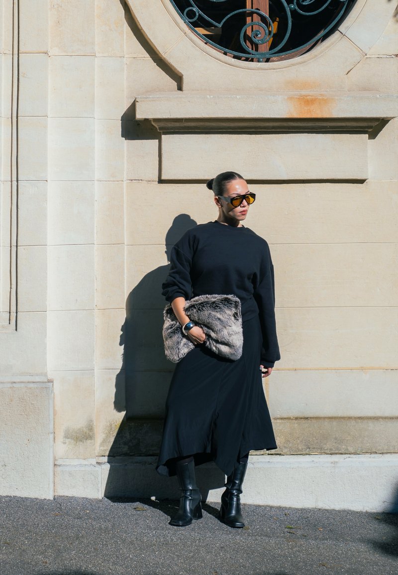 Frau in schwarzem Kleid und Stiefeln steht gegen eine beige Steinmauer und hält eine große graue Fell-Clutch in der Hand, während sie getönte Sonnenbrillen trägt.