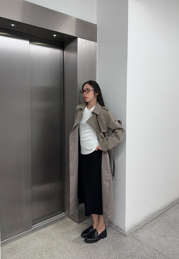 Une femme portant des lunettes et un long manteau à carreaux attend près d’un ascenseur fermé dans un bâtiment moderne.