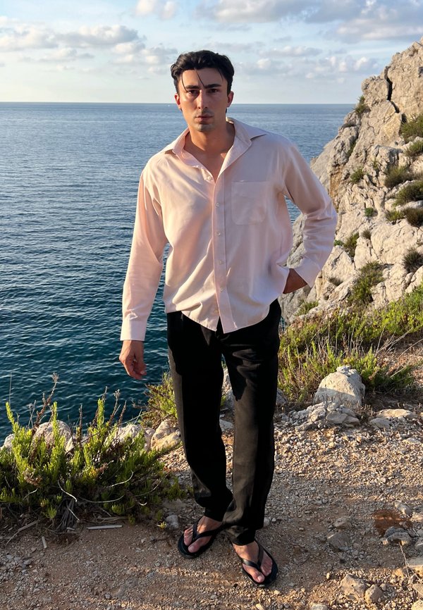 Jeune homme debout au bord d'une falaise rocheuse près de la mer, portant une chemise rose clair, un pantalon noir et des tongs, la main dans la poche et une expression sérieuse.