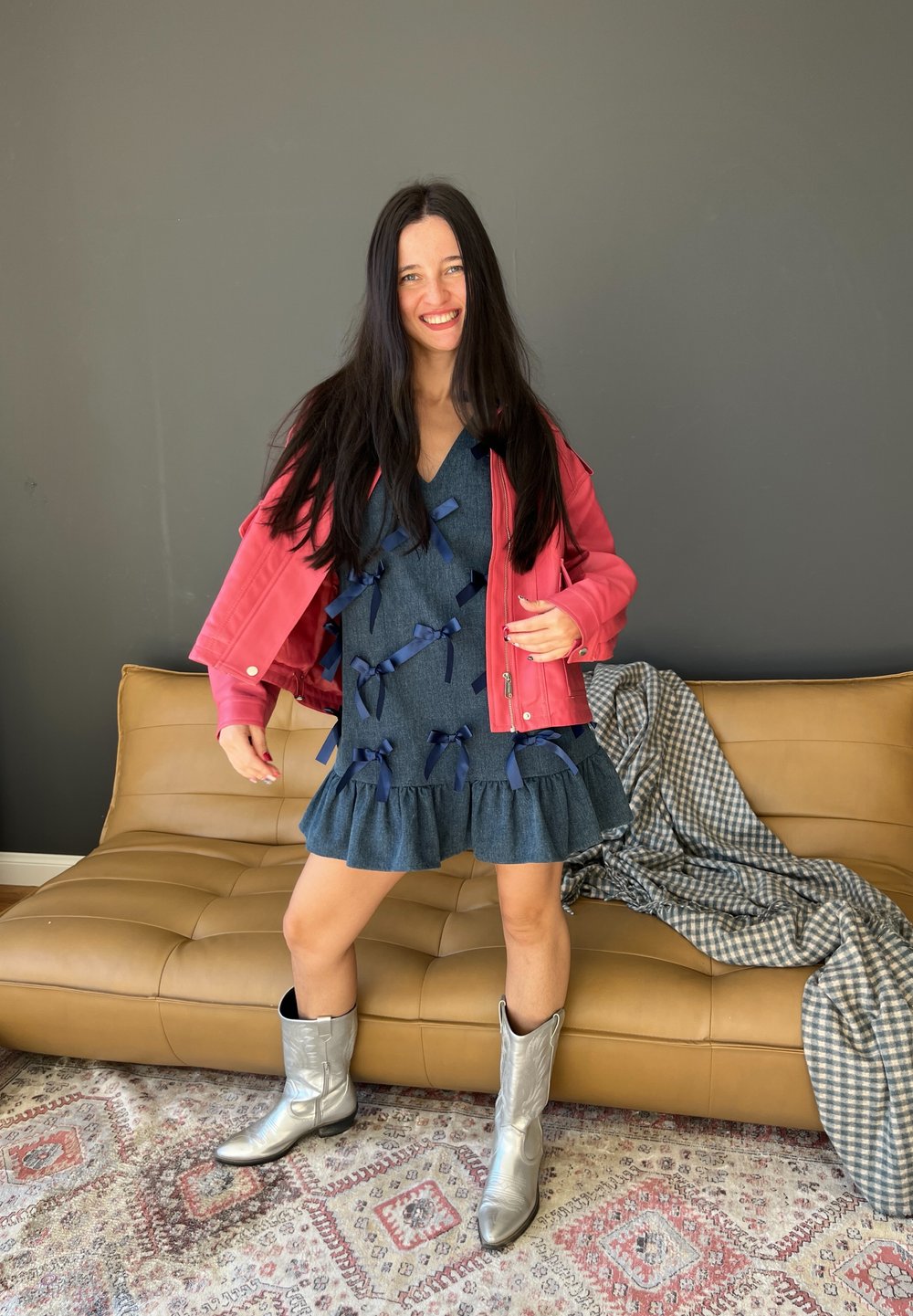 Chaqueta de cuero rosa sobre un vestido de mezclilla con detalles en cinta azul y dobladillo con volantes, combinado con botas vaqueras plateadas brillantes.