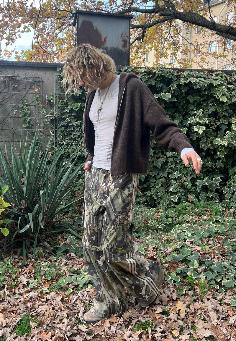 Brown zip-up hoodie, white fitted shirt, camo cargo trousers, and beige shoes. Background features greenery and fallen leaves.