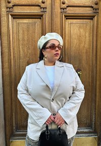 Sacou oversized bej cu mâneci lungi, purtat peste un top alb. Beretă albă și ochelari de soare negri. Ține o poșetă mică neagră.