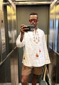 White tunic featuring orange embroidery and tassels, matched with brown shorts. Wearing sunglasses and holding a green bag in a mirrored lift.