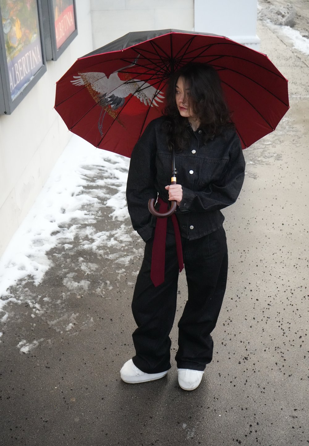 Femme en veste et pantalon noirs tenant un parapluie rouge avec un motif d'oiseau, debout sur un trottoir mouillé avec des plaques de neige à proximité.