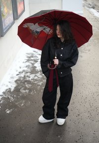 Femme en veste et pantalon noirs tenant un parapluie rouge avec un motif d'oiseau, debout sur un trottoir mouillé avec des plaques de neige à proximité.