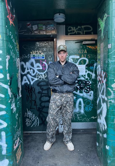 Pantalones de camuflaje y una chaqueta bomber oscura, complementados con una gorra de camuflaje. El fondo presenta paredes con muchas pintadas y una puerta.