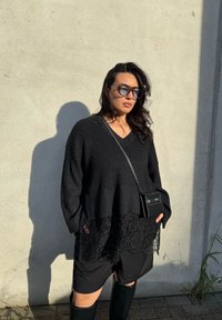 Frau trägt übergroßen schwarzen Strickpullover mit Spitzenabschluss, schwarze Sonnenbrille, Umhängetasche, kniehohe Stiefel, steht vor einer sonnenbeschienenen Betonwand.