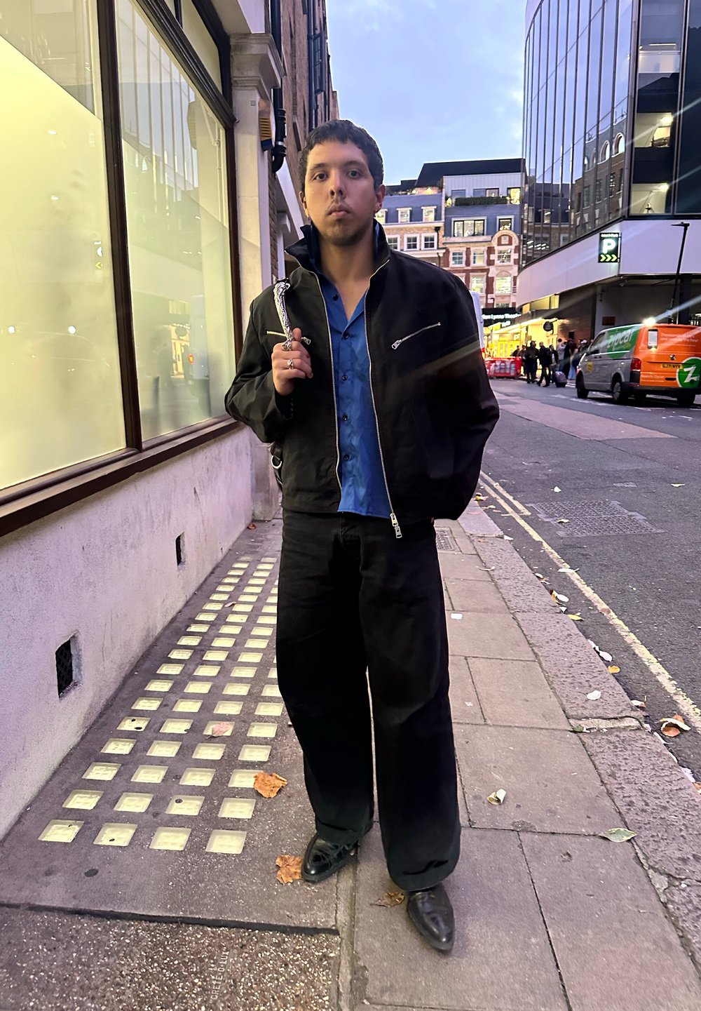 Must mustriga bomberjakk, all sinine särk, laiad mustad püksid ja mustad kingad, seisab linna kõnniteel klaasakende lähedal.