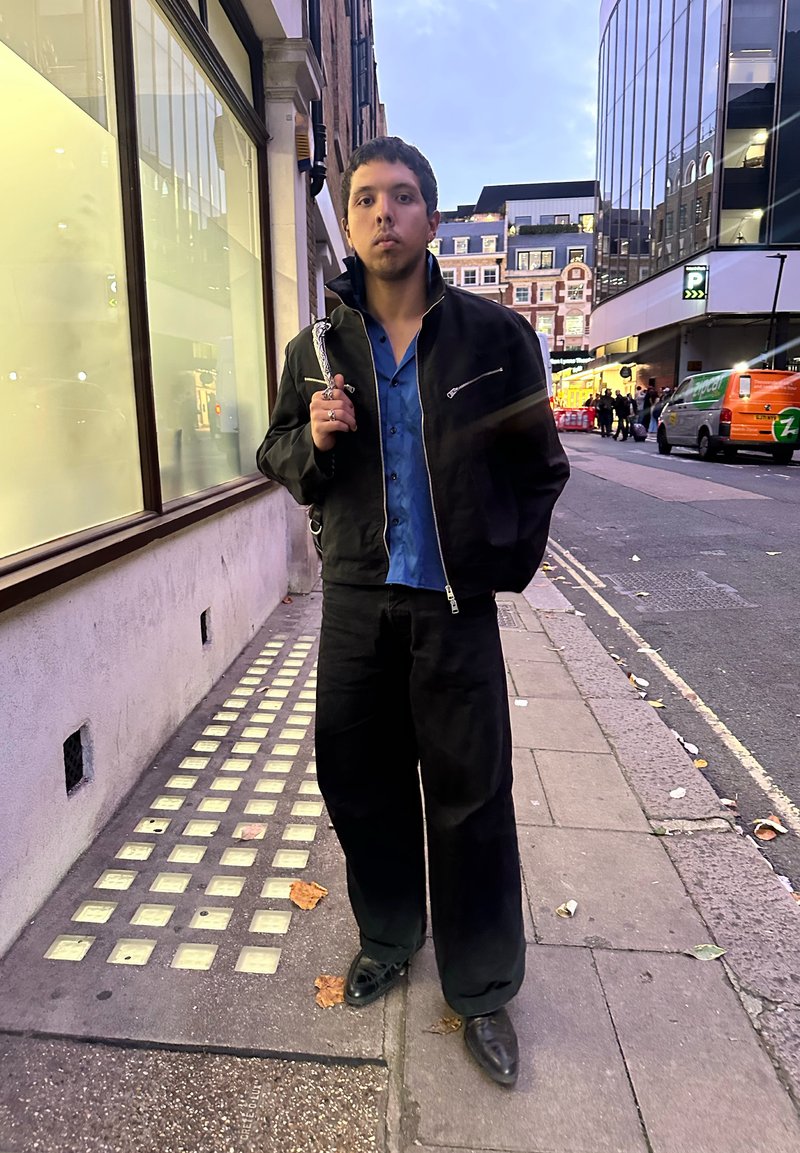 Black bomber jacket with zip, blue shirt underneath, loose black trousers, and black shoes, standing on a city pavement near glass windows.