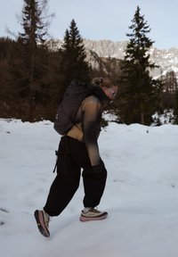 Personne portant des lunettes de soleil, un sac à dos et des vêtements de randonnée marchant en montée sur la neige avec des sapins et des sommets de montagnes en arrière-plan.