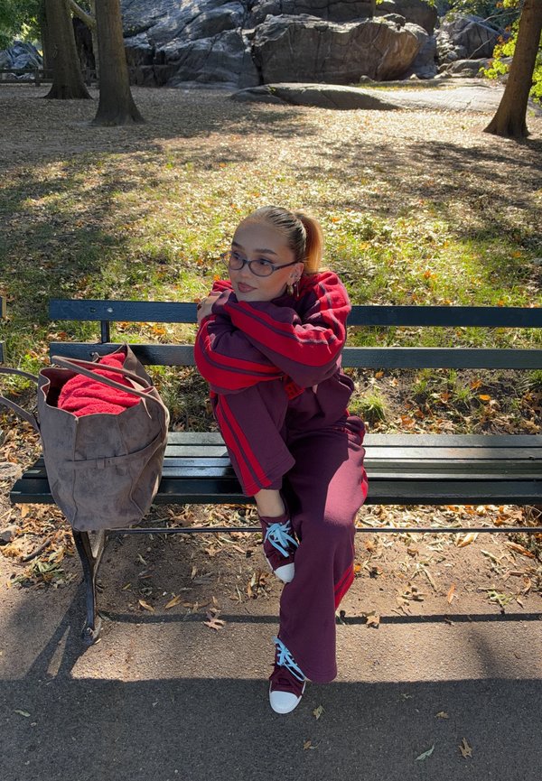 Una mujer con un chándal rojo y gafas está sentada en un banco del parque junto a una bolsa de tela, con árboles y afloramientos rocosos detrás.