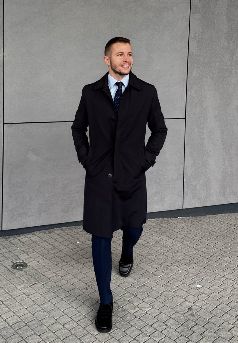 Man in black coat, navy suit, and tie walking on grey tiled pavement with hands in pockets, smiling and looking to the right.