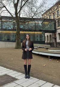 Γυναίκα με σκούρο παλτό, γκρι πουλόβερ, φούστα και μαύρες μπότες που στέκεται σε μονοπάτι έξω κοντά σε ένα μεγάλο δέντρο και μοντέρνο γυάλινο κτίριο το φθινόπωρο.