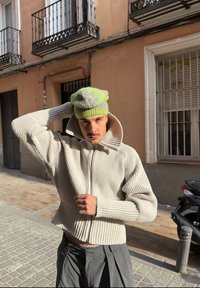 Jeune homme portant un pull beige côtelé et un bonnet vert ajuste son chapeau en se tenant dans une rue ensoleillée avec des balcons et un scooter à proximité.
