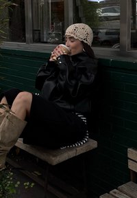 Casaco oversized de couro preto, vestido preto com bainha riscada, chapéu de crochet bege, a beber de um copo de papel enquanto está sentado num banco de madeira.