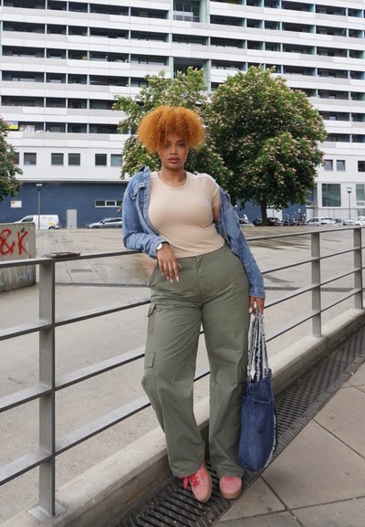 Camisa ajustada beige debajo de una chaqueta de denim clara, pantalones cargo verdes oliva, zapatos rosa y una bolsa de denim azul. Entorno urbano con vegetación.