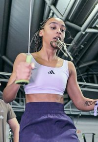 Figura atlética saltando la cuerda, vistiendo un top corto morado claro con un logo negro y pantalones cortos oscuros, destacando los músculos de los brazos y las trenzas.