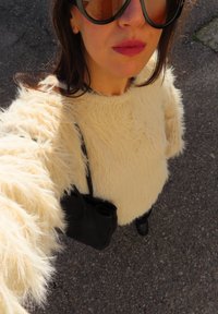 Krämfärgad fluffig tröja med långa ärmar, med en texturerad, lurvig yta. En svart handväska syns, tillsammans med svarta byxor och skor.