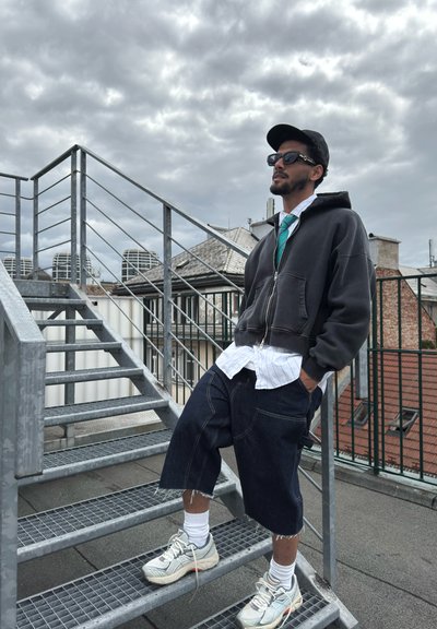 Sudadera gris con cremallera, camisa blanca con cuello, shorts de mezclilla oscuros hasta la rodilla, calcetines blancos y zapatillas grises; de pie en unas escaleras de metal con cielo nublado.