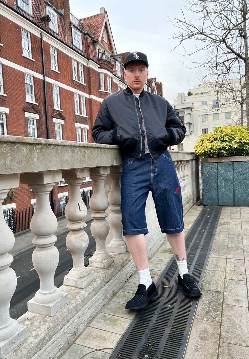 Μαύρο μπουφάν, γκρι πουκάμισο, τζιν σορτσάκι με κόκκινη κεντητική, λευκές κάλτσες και μαύρα παπούτσια. Φόντο με πέτρινη περίφραξη και πλακόστρωτο διάδρομο.