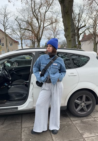 Chaqueta de mezclilla azul claro sobre un cuello de tortuga oscuro, combinada con pantalones de chándal grises anchos y botas negras. Un gorro azul completa el look. Coche cerca.