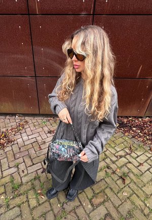 Suéter gris de punto con cable, pantalones negros con rayas diplomáticas y un bolso colorido con estampado y correa para el hombro, sobre un fondo de pavimento de ladrillos.