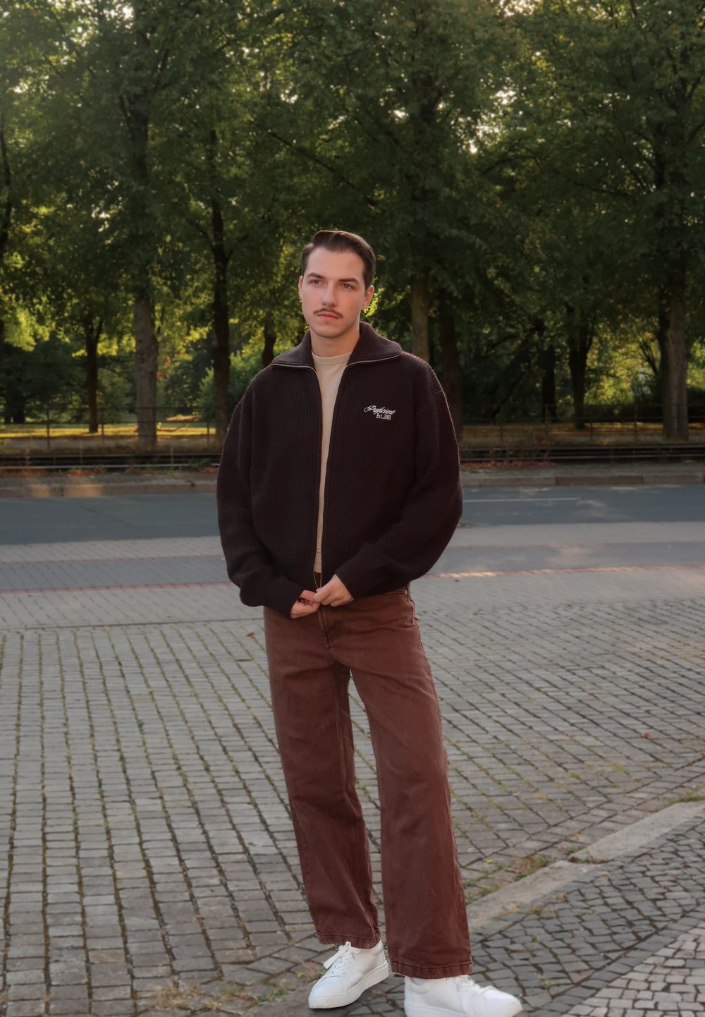 Männerjacke in Dunkelbraun, mit Reißverschluss und Strickmuster, beiges T-Shirt, braune Hose und weiße Sneakers. Steht auf Kopfsteinpflaster, umgeben von Grün.