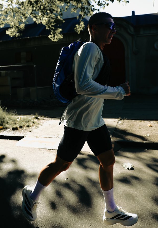 Homme portant des lunettes de soleil et un sac à dos bleu fait du jogging en plein air par une journée ensoleillée avec des ombres sur le trottoir.
