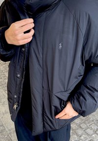 Melnā polsterētā jaka ar rāvējslēdzēja aizdari un sprādzēm, ar nelielu izšūtu logo uz krūtīm un fakturētu audumu.