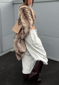 Femme portant un manteau en fourrure marron sur un pantalon de survêtement gris court et des bottes hautes marron foncé, tenant un sac à main beige à motif.