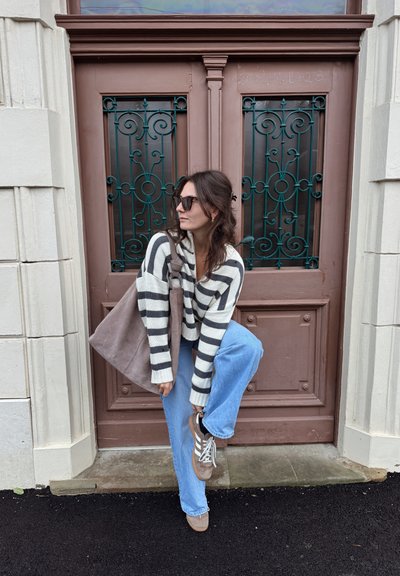 Suéter a rayas en crema y gris, jeans azul claro, bolso topo y zapatillas con rayas de acento. Puerta marrón con trabajo en metal decorativo.