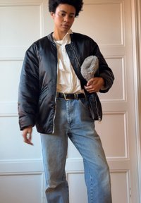 Μαύρο επενδυμένο bomber μπουφάν με φερμουάρ μπροστά, φορεμένο πάνω από λευκό πουκάμισο, σε συνδυασμό με ανοιχτό μπλε ψηλόμεσα τζιν. Ασημένια κοσμήματα ως αξεσουάρ.