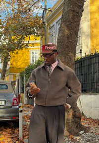 Veste marron à motif pied-de-poule, chemise rose à col, cravate à motif. Porte une casquette marron avec le texte "Project". Tient un smartphone en plein air.