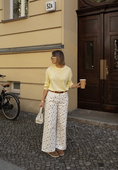 Suéter amarillo texturizado, pantalones anchos blancos con estampado floral, zapatos beige, sosteniendo una taza de café y un bolso crema. Entorno natural al aire libre.
