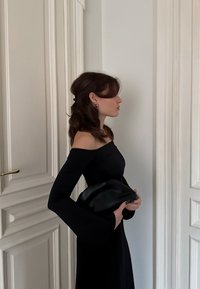 Femme aux cheveux bruns bouclés portant une robe noire épaules dénudées, tenant un sac à main noir, debout de profil près de portes blanches à panneaux.