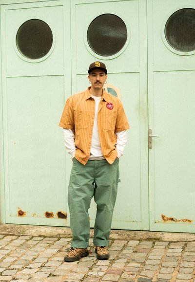 Camisa de manga corta a rayas marrones sobre una camisa de manga larga blanca, pantalones verdes holgados y zapatos de piel de leopardo, de pie frente a una puerta verde menta.