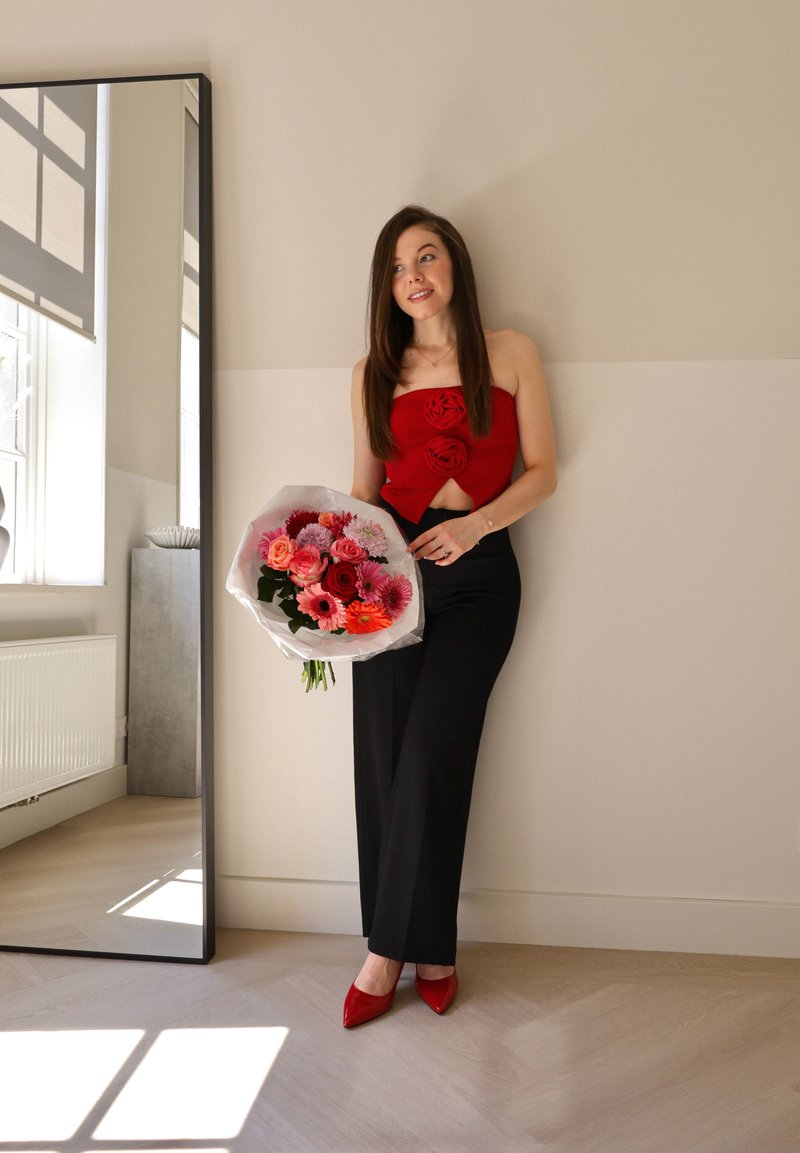 Κόκκινο τοπ χωρίς τιράντες με λουλουδάτες λεπτομέρειες, μαύρο παντελόνι και κόκκινα μυτερά παπούτσια. Κρατώντας μια ανθοδέσμη με ροζ, κόκκινα και μωβ λουλούδια σε διαφανή περιτύλιξη.