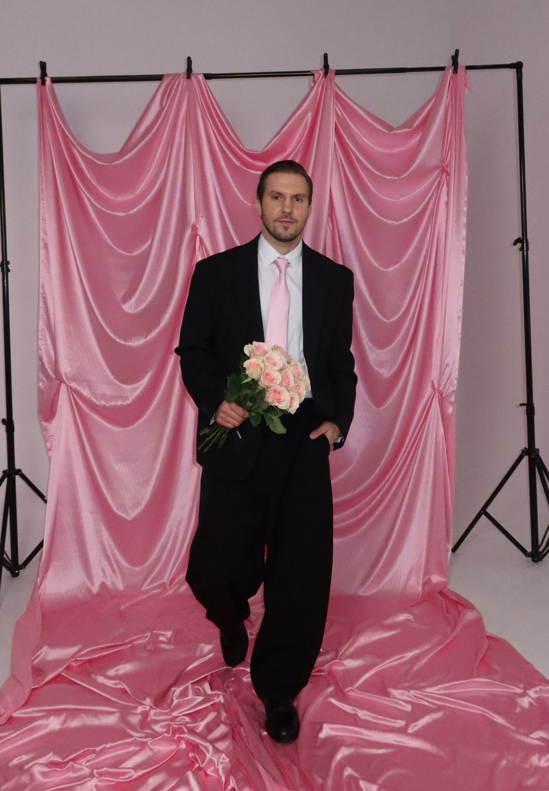 Homme en costume noir et cravate rose tenant des roses roses, debout sur et devant un fond de tissu rose brillant drapé.