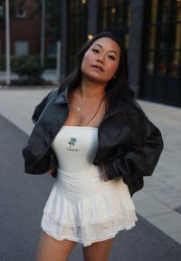 Čierna skrátená kožená bunda cez biele šaty bez rukávov s vrstvenou volánovou sukňou. Šaty majú vyšívané modré ruže a text.