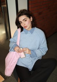 Suéter azul claro sobre uma camisa quadrada, combinado com calças pretas. Bolsa de fleece rosa segurada nas mãos, exibindo anéis nos dedos. Superfícies texturizadas visíveis.