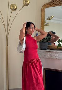 Robe rouge sans manches avec corsage ajusté et ourlet évasé. Le modèle tient un sac à main argenté métallique. En arrière-plan, un mur clair et une lampe sur pied.