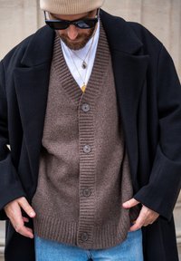 Cardigan en tricot brun avec col en V, ourlet côtelé et cinq boutons, superposé sous un manteau noir et porté sur un T-shirt blanc.