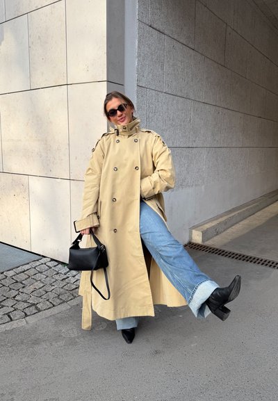 Mujer con gabardina beige, gafas de sol, vaqueros azules y botas negras posando al aire libre con una pierna levantada, sosteniendo un bolso negro.