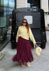 Femme portant des lunettes de soleil, un pull jaune et une jupe bordeaux superposée, marchant devant un véhicule vintage noir, tenant un sac à main beige.