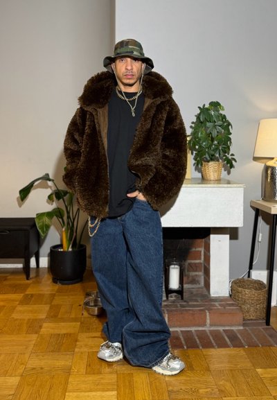 Hombre con abrigo marrón de piel sintética, camisa negra, vaqueros anchos azules, zapatillas deportivas y un sombrero tipo bucket de camuflaje, de pie en el interior cerca de plantas y una chimenea.