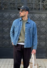 Denim jakna v modri barvi z dvema žepoma na prsih, prekrivajoča zeleni pulover, v kombinaciji s črnimi hlačami in bež torbo.