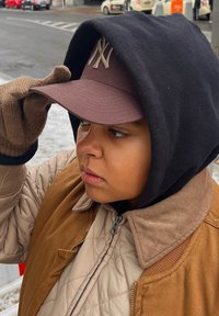 Άτομο φορώντας καπέλο μπέιζμπολ καφέ, μαύρη φούτερ με κουκούλα και καφετί γεωμετρικό μπουφάν. Τα γάντια είναι καφέ και έχουν πλεχτή υφή. Έκφραση προσώπου επικεντρωμένη.