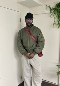 Homme portant une veste bomber vert olive, un pantalon de survêtement gris, un bonnet noir, des lunettes et un sac bandoulière rouge, debout à l'intérieur près de murs blancs avec des plantes.