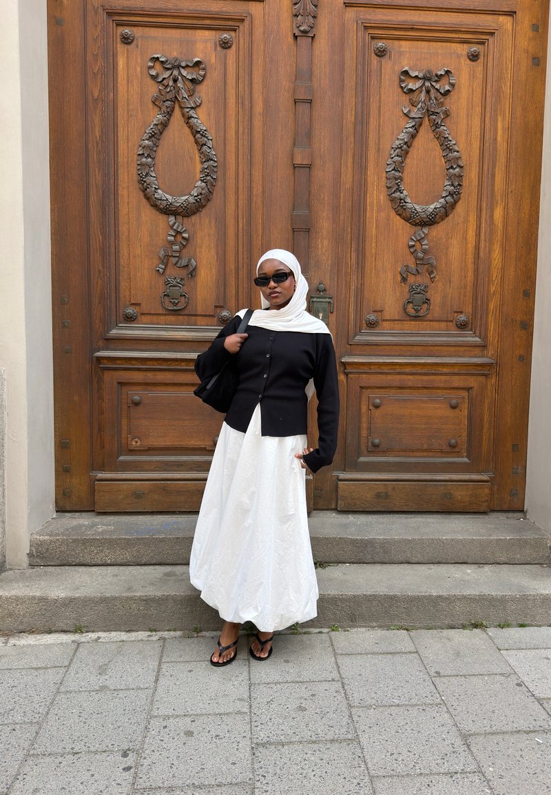 Femme portant un hijab blanc, un cardigan noir et une jupe blanche debout sur des marches devant de grandes portes en bois avec des décorations de couronnes sculptées.